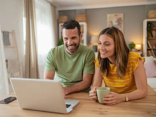 A couple smiling at a laptop at their kitchen table